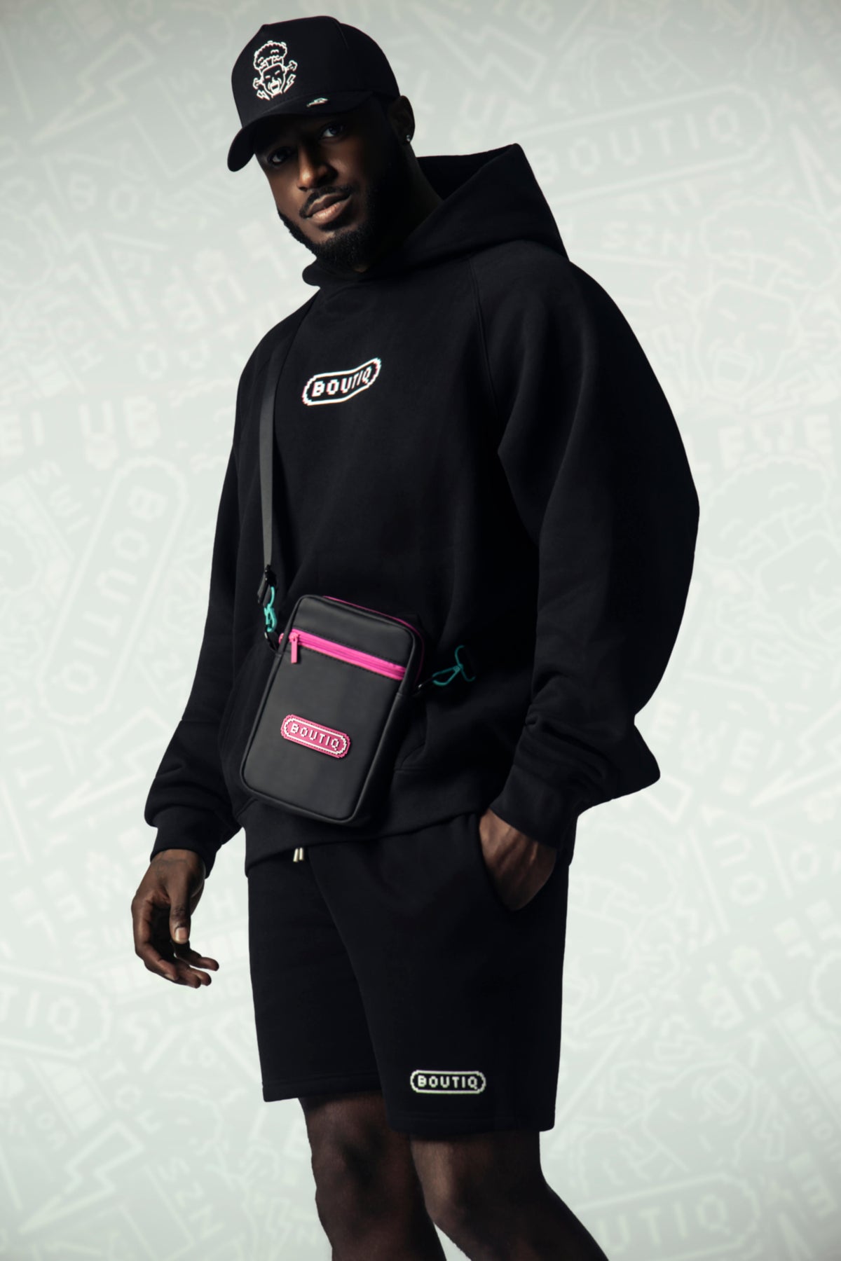 Model in a black BOUTIQ hoodie, shorts and snapback with a pink-trim crossbody, face fully visible.
